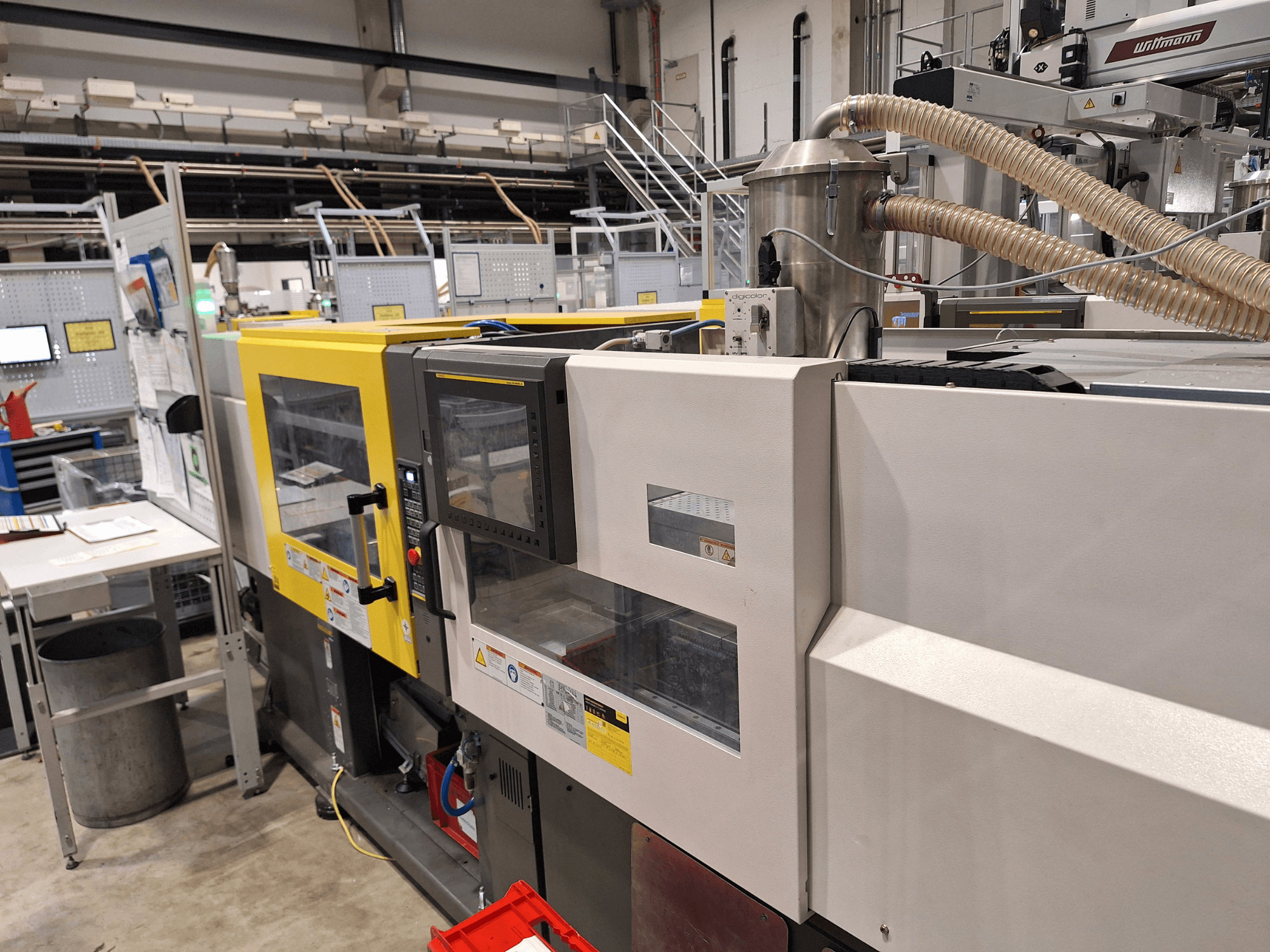 Front view of a multi-colored injection molding machine, featuring yellow and gray sections, with control panel and exhaust system.