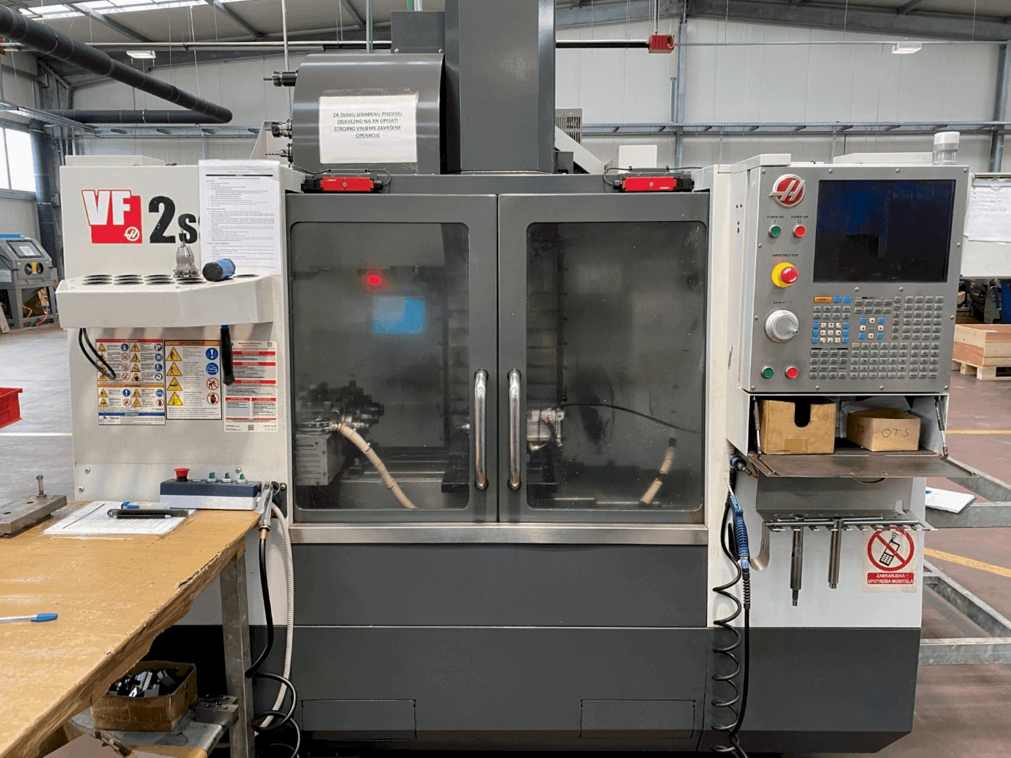 Haas VF-2SS machining center in front view, featuring control panel, coolant hose, and safety labels on the side.